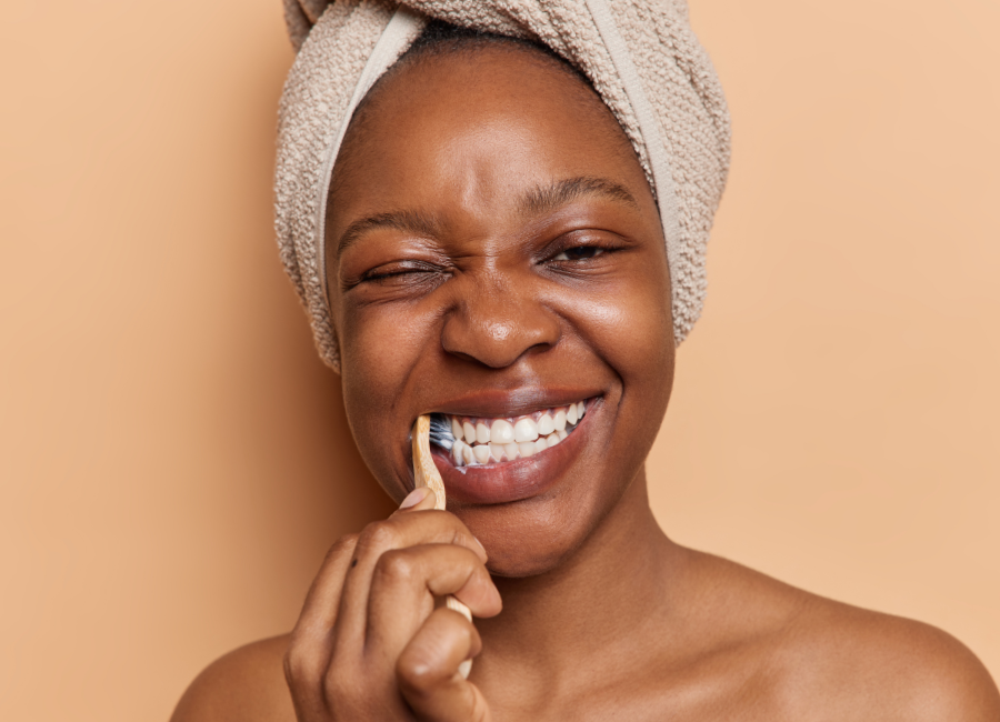 Woman with hair in towel smiling broadly while brushing teeth