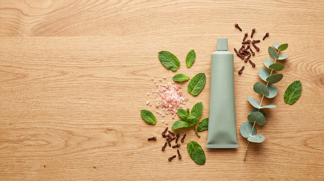 Botanical flat lay of a natural toothpaste tube surrounded by peppermint, pink salt, clove and eucalyptus on oak — Best Natural Toothpaste in Australia 2026.