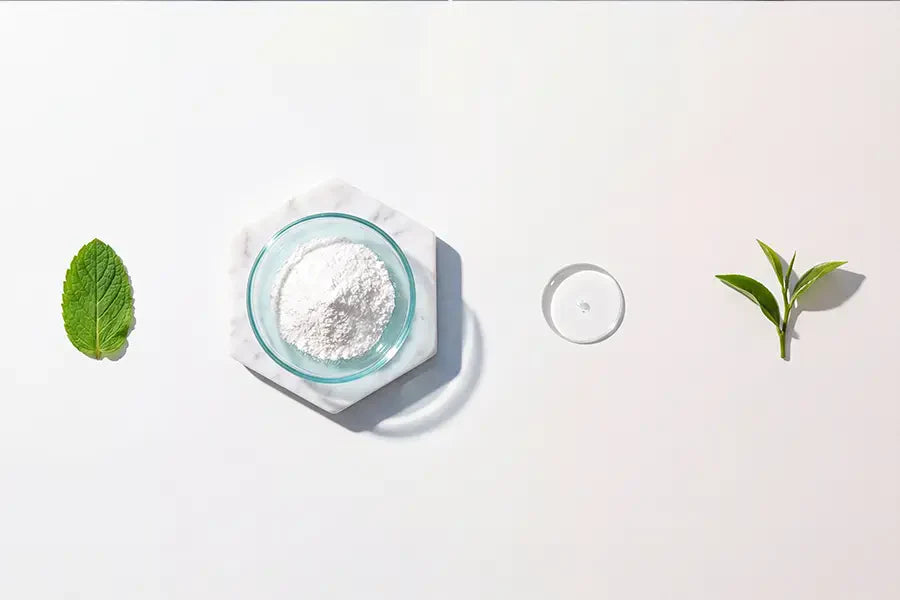 Minimal arrangement of clean toothpaste ingredients — mint leaf, mineral powder, oil droplet, and green tea