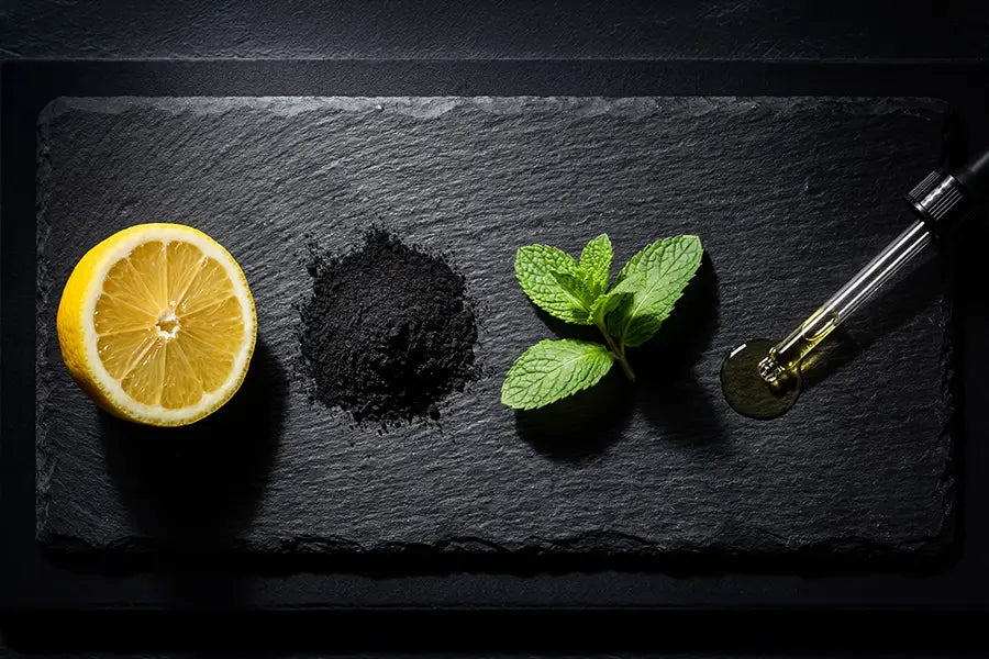 Editorial flat lay of natural whitening ingredients — charcoal powder, lemon, mint, and oil droplet on dark slate surface