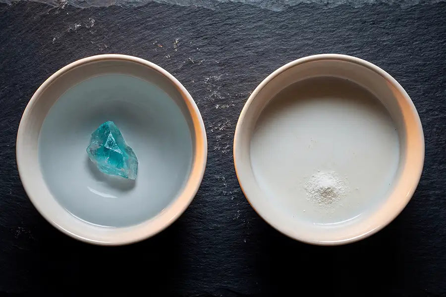 Two ceramic bowls on dark slate — aqua mineral water and white mineral powder side by side representing the hydroxyapatite vs fluoride comparison