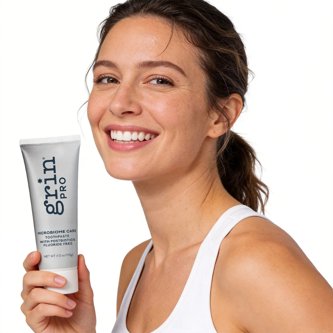 Smiling person holding a tube of Grin Pro Microbiome Care fluoride-free toothpaste against a white background