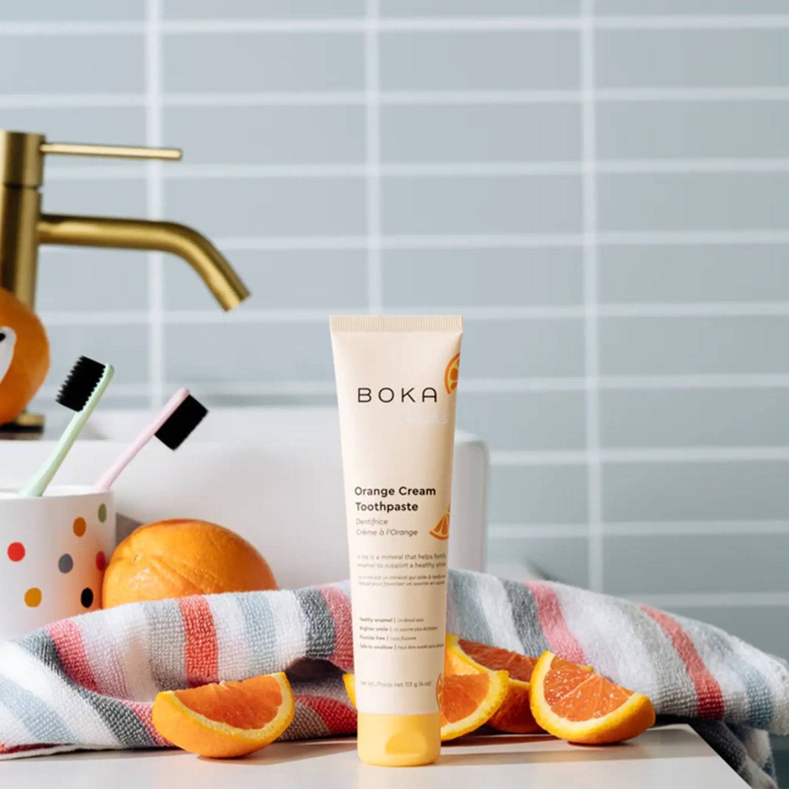 Boka Kids Orange Cream toothpaste on a bathroom bench with fresh oranges, a striped towel and toothbrushes