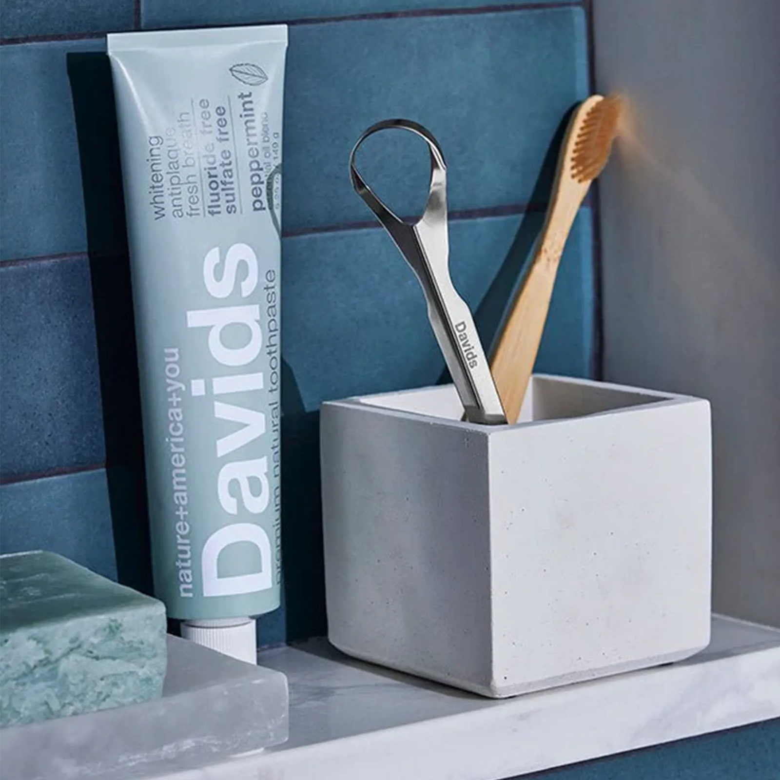 Davids stainless steel tongue scraper in a bathroom holder with a Davids toothpaste tube and bamboo toothbrush