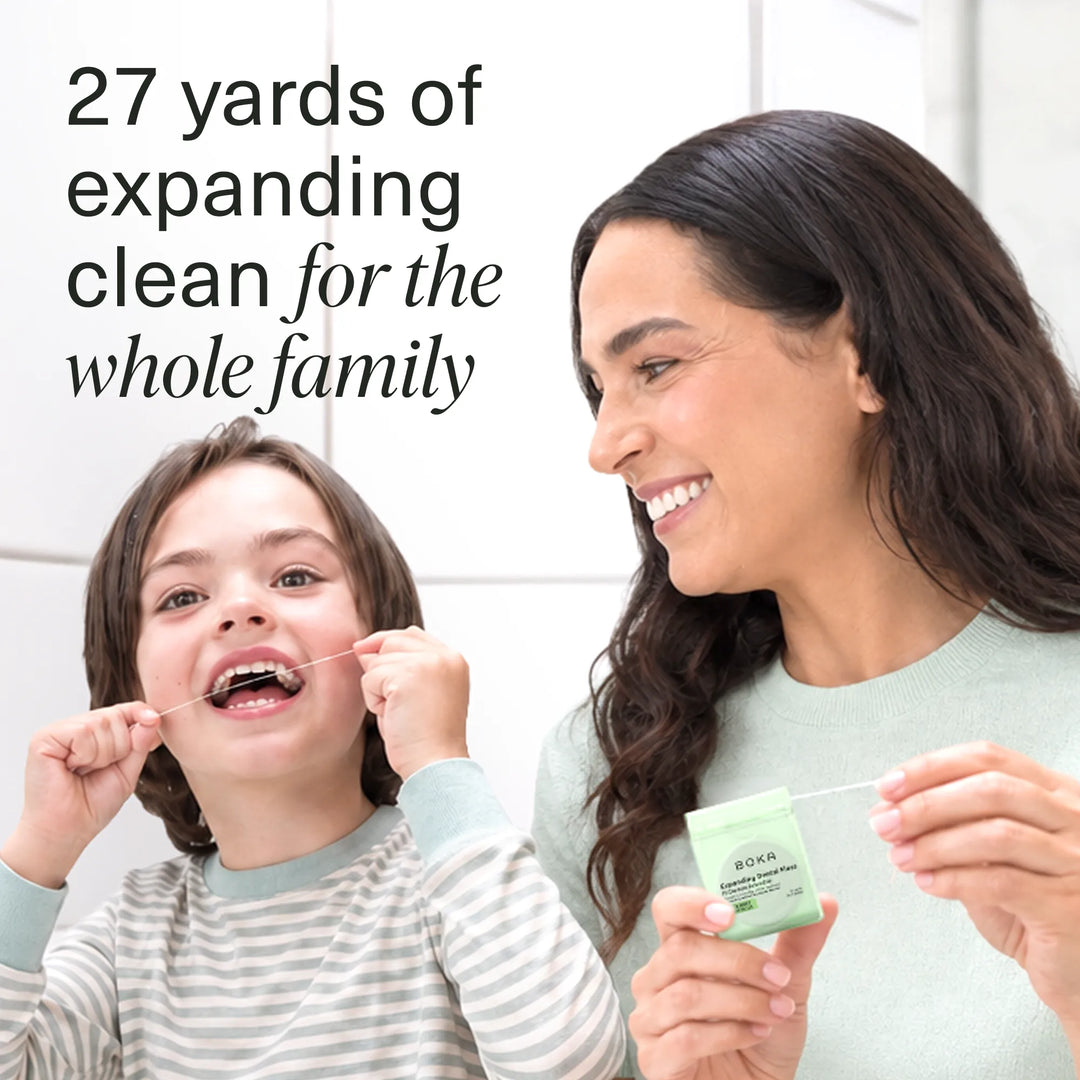 Smiling child flossing while a woman holds the green Boka Expanding Dental Floss dispenser in a white tiled bathroom