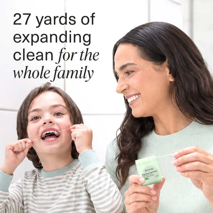 Smiling child flossing while a woman holds the green Boka Expanding Dental Floss dispenser in a white tiled bathroom