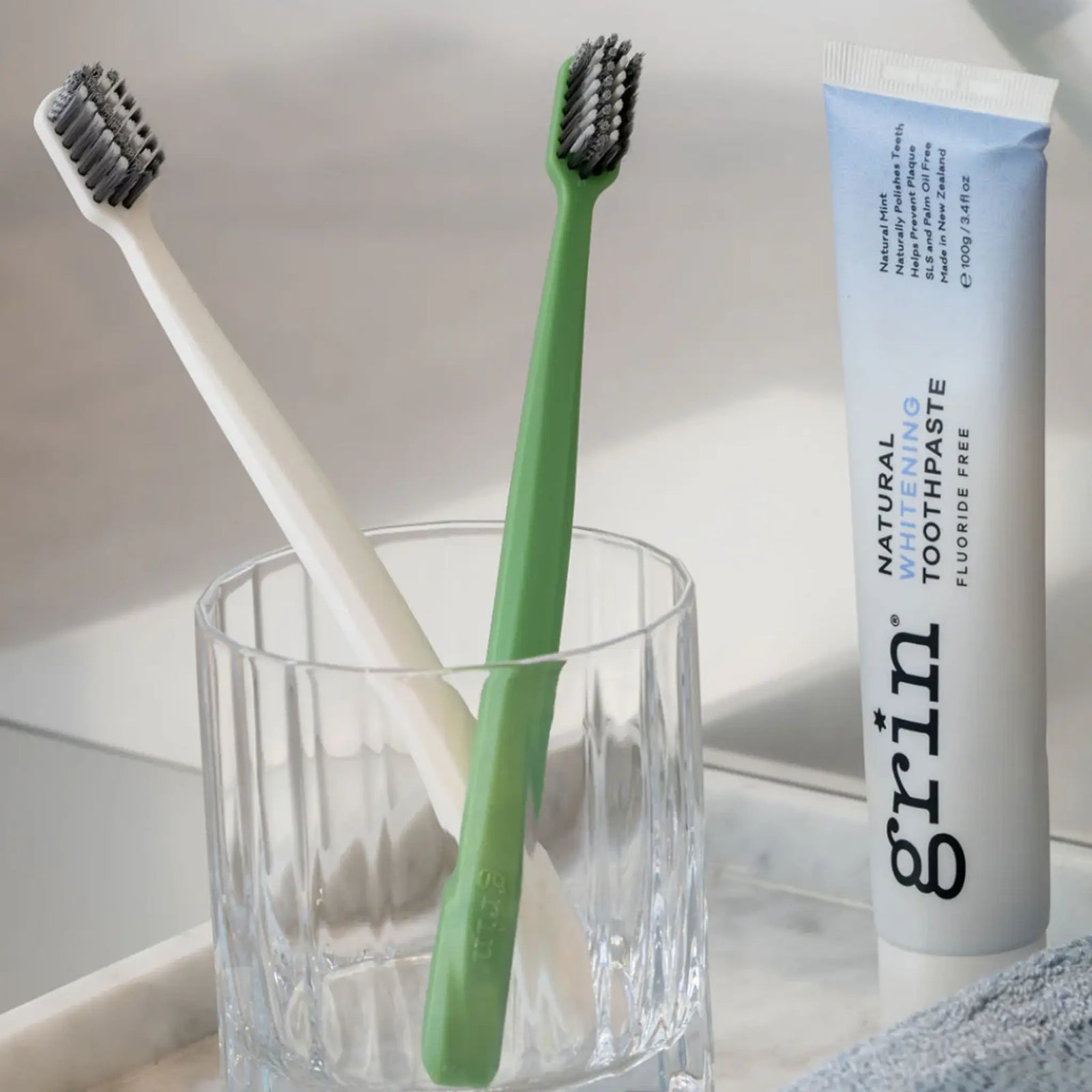 Two toothbrushes in a glass with a tube of grin toothpaste on a light surface.