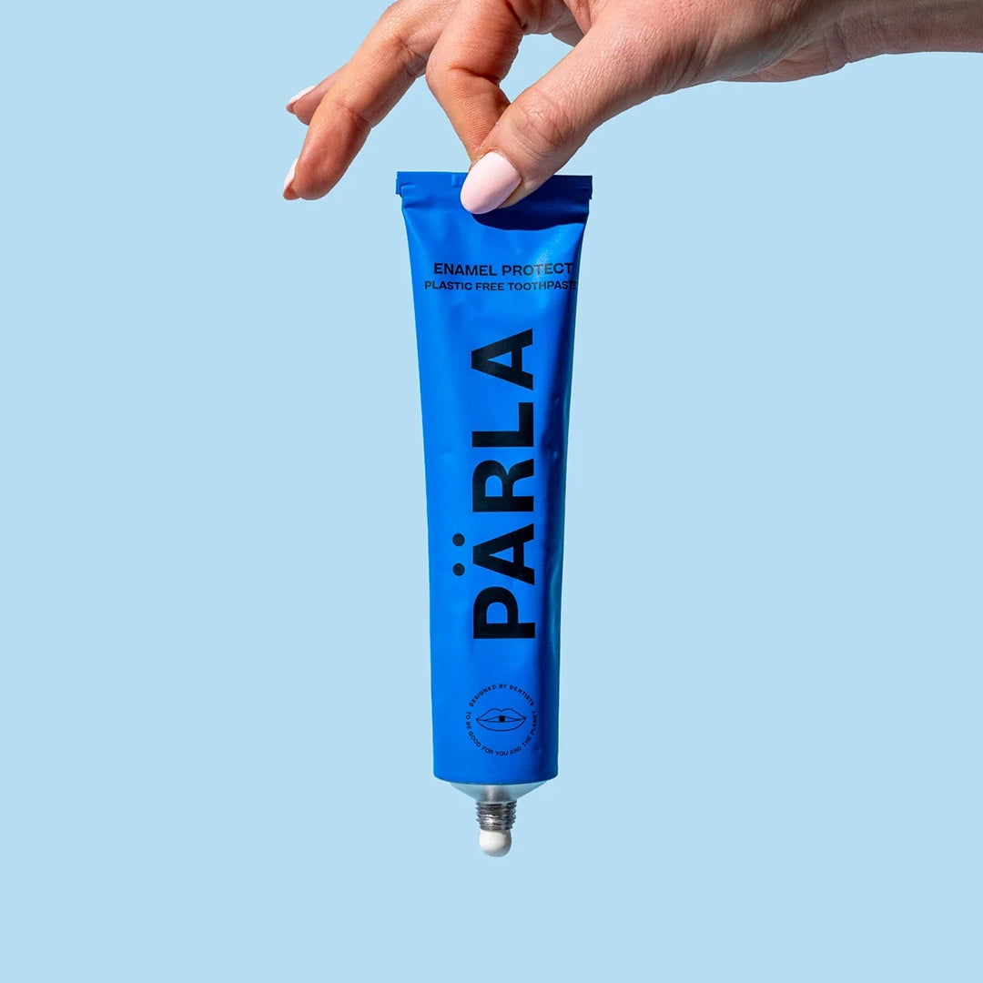 Hand holding a blue PÄRLA Enamel Protect plastic free toothpaste tube against a pale blue background