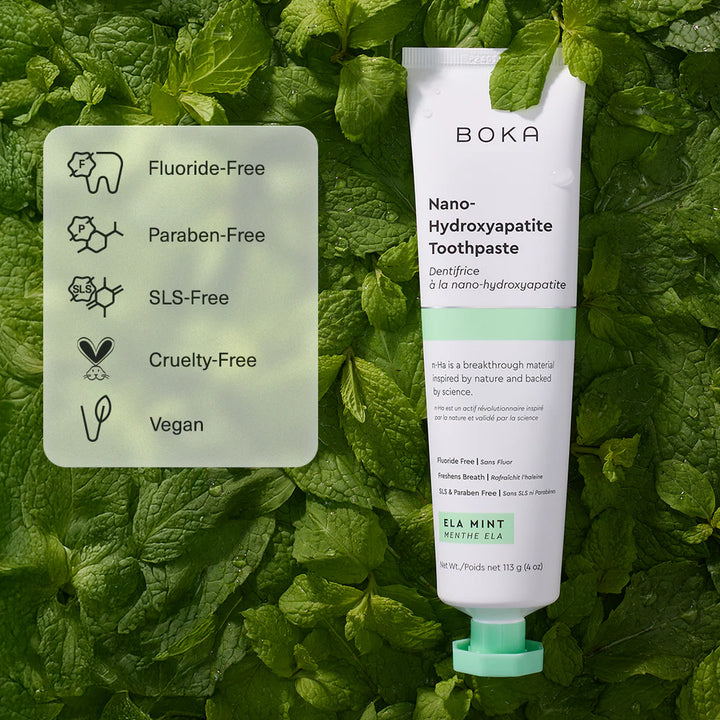 A tube of Boka Nano-Hydroxyapatite Toothpaste in mint flavor is displayed against a background of fresh green mint leaves, next to a translucent square card detailing product features like &