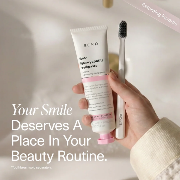 Hand holding a Boka Cherry Blossom toothpaste tube and a Boka toothbrush at a white basin